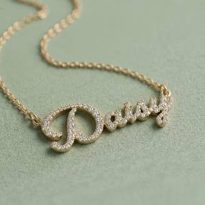 Könnte beinhalten: Eine goldfarbene Halskette mit einem Namensanhänger in Schreibschrift, der den Namen Daisy bildet. Der Anhänger ist auf der gesamten Oberfläche der Buchstaben mit zahlreichen kleinen, klaren, funkelnden Steinen besetzt und an einer dünnen Kette befestigt.