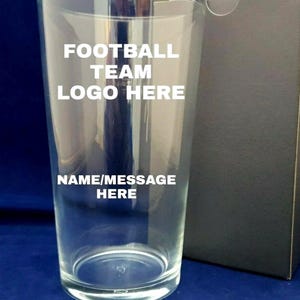 May include: A clear pint glass with the text "FOOTBALL TEAM LOGO HERE" and "NAME/MESSAGE HERE" in white. The glass is next to a black box on a blue surface.