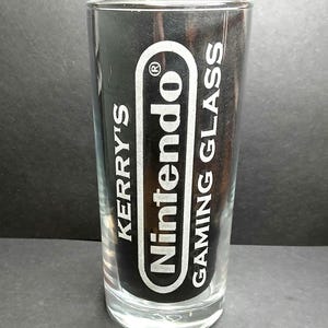 May include: A clear glass tumbler featuring white printed text that reads KERRY'S Nintendo GAMING GLASS. The Nintendo logo is enclosed within a white oval outline. The glass has a cylindrical shape with a thick, transparent base and smooth walls.