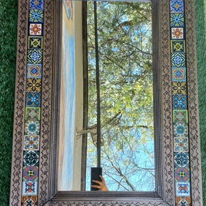 May include: Rectangular decorative mirror with a mosaic tile border. The border features colorful square tiles with intricate patterns in shades of blue, red, yellow, and green. The frame is a textured bronze color. The mirror reflects a view of a tree and sky.