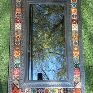 May include: Rectangular mirror with a decorative, patterned border. The border features colorful square tiles in various designs and colors, set against a textured, silver-toned frame. The mirror reflects a tree and sky.