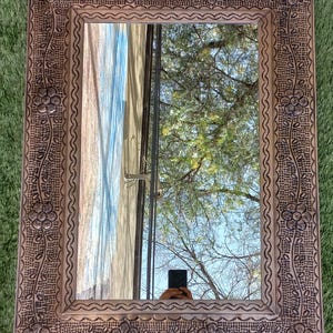 May include: Rectangular mirror with a decorative, embossed copper-colored frame. The frame features floral and vine patterns, with a scalloped edge. The mirror reflects a view of trees and sky. The mirror is a decorative home decor item.