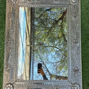 May include: A rectangular silver mirror with a detailed floral and dotted pattern frame. The mirror reflects a tree and sky. The frame has a raised floral design at each corner and along the sides. The mirror is on a green surface.