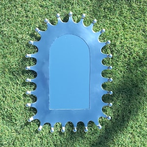 Puede incluir: Un espejo plateado con un marco festoneado único. El marco tiene una serie de bordes curvos, cada uno coronado con una pequeña bola plateada redonda. El espejo es rectangular con esquinas superiores redondeadas, reflejando un tono azul.