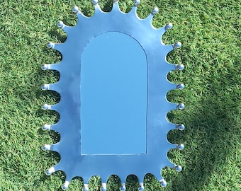 May include: A silver mirror with a distinctive scalloped frame. The frame features a series of curved edges, each with a small, round silver ball. The mirror has a rectangular shape with rounded top corners, reflecting a blue colour.