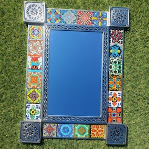 May include: Rectangular mirror with a blue frame and colorful, patterned tiles. The tiles feature various geometric and floral designs in shades of red, orange, yellow, and green. The mirror is surrounded by a dark blue frame with a wavy edge.