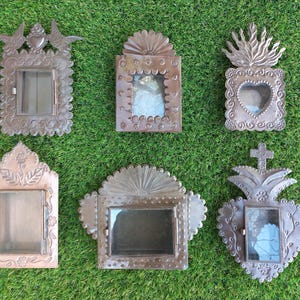 May include: Six handcrafted, rustic metal frames with glass fronts, arranged on a green surface. The frames feature various decorative elements, including birds, hearts, and floral designs. Each frame has a hinged door.