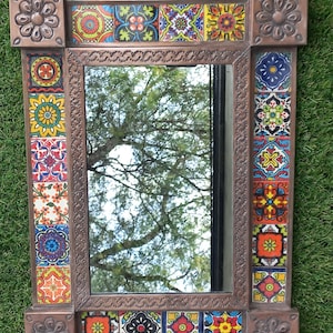 May include: Rectangular mirror with a decorative frame. The frame is a dark bronze color and features colorful, patterned tiles. The corners have embossed floral designs. The mirror reflects a tree and sky.