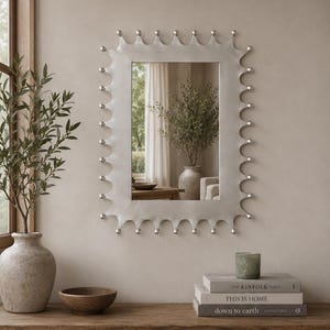 Puede incluir: Un espejo de pared rectangular con un marco de tono plateado que presenta un borde festoneado adornado con pequeños acentos metálicos esféricos a lo largo de todo el perímetro. La superficie de vidrio reflectante muestra una vista clara.