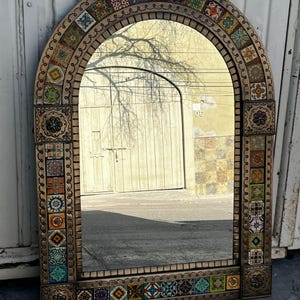 May include: An arched mirror with a decorative frame. The frame is adorned with colorful, patterned tiles and a border of small, repeating designs. The mirror reflects a street scene.