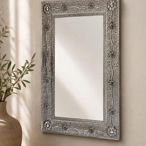 May include: A rectangular wall mirror featuring a silver-toned frame with an intricate embossed pattern. The frame displays a repeating floral motif, wavy lines, and a textured dotted background, with larger flower accents at each corner.