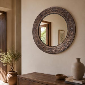 May include: A round wall mirror featuring a wide, dark bronze-toned frame with an intricate embossed pattern of floral motifs and textured dots. The circular glass surface reflects the surrounding room, including a doorway and a framed picture.