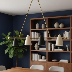 Op de afbeelding: Een moderne, messingkleurige kroonluchter met drie kegelvormige lampen in zwart en crème. De armatuur hangt boven een houten eettafel, met een donkerblauwe muur en een boekenkast op de achtergrond. De kamer is ingericht met grijze stoelen en een grote potplant.