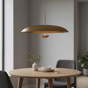 Könnte beinhalten: Eine moderne, goldfarbene Pendelleuchte hängt über einem runden Esstisch aus Holz. Die Leuchte hat einen großen, flachen Schirm und ein kleineres, nach unten gerichtetes Licht. Eine kleine Vase mit Grün und ein Gedeck stehen auf dem Tisch.