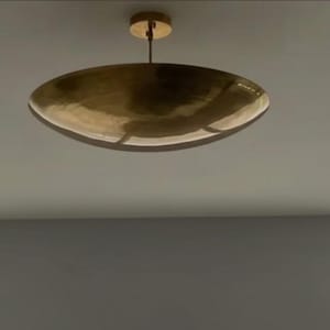 Puede incluir: Una lámpara de techo dorada de forma ovalada. La luz está suspendida de un dosel circular por una fina varilla. La lámpara tiene una superficie reflectante y el diseño general es minimalista y moderno.