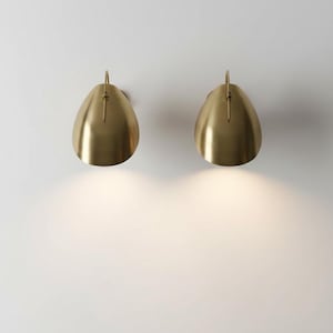 Könnte beinhalten: Zwei goldfarbene Wandleuchten in Tropfenform mit einem kleinen, gebogenen Griff oben. Die Leuchten sind an einer weißen Wand befestigt, und das Licht der Leuchten beleuchtet die Wand darunter.