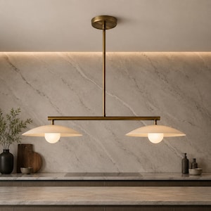 May include: A linear pendant light fixture featuring a central brass-toned rod and horizontal bar supporting two wide, shallow, matte white saucer-shaped shades. Each shade contains a central glowing spherical bulb, suspended from the ceiling.