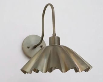 May include: A wall-mounted light fixture with a ruffled, bell-shaped shade. The fixture has a curved arm and a circular base, all in a muted brass finish. The shade has a scalloped edge, adding a decorative touch to the design.
