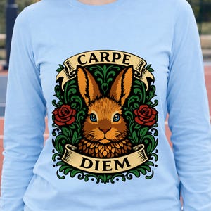 May include: Light blue long-sleeve shirt featuring a detailed illustration. The design includes a brown rabbit with blue eyes, surrounded by green foliage and red roses. The words "CARPE" and "DIEM" are displayed on banners.
