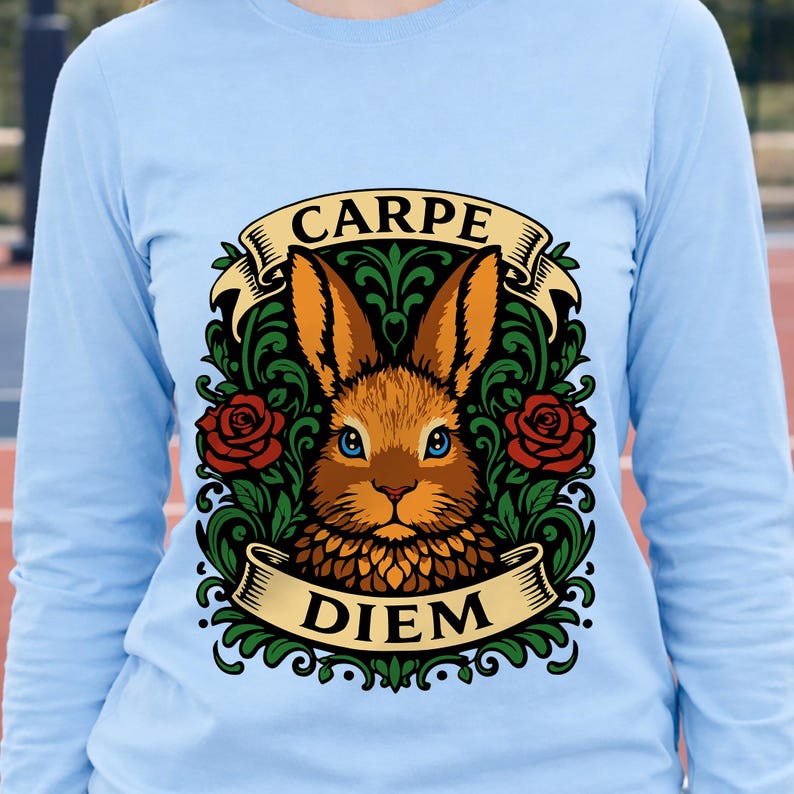 May include: Light blue long-sleeve shirt featuring a detailed illustration. The design includes a brown rabbit with blue eyes, surrounded by green foliage and red roses. The words "CARPE" and "DIEM" are displayed on banners.