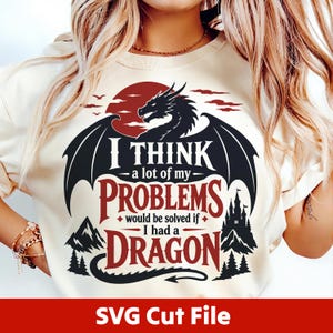 Op de afbeelding: Een crèmekleurig t-shirt met het silhouet van een zwarte draak tegen een rode zon, met de tekst "I THINK a lot of my PROBLEMS would be solved if I had a DRAGON" in zwart en rood, naast illustraties van een kasteel en bergen.