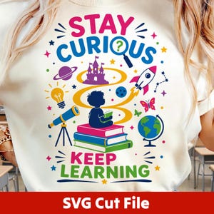 May include: A cream-colored t-shirt featuring a colorful graphic with the text STAY CURIOUS and KEEP LEARNING. The design includes a child sitting on a stack of books, a rocket, a telescope, a globe, a lightbulb, a castle, and a planet.