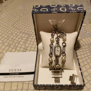May include: A Guess watch with a silver bracelet and a white watch face, presented in a branded box. The watch has pearl accents and is displayed on a white pillow. The box and warranty card are also visible.
