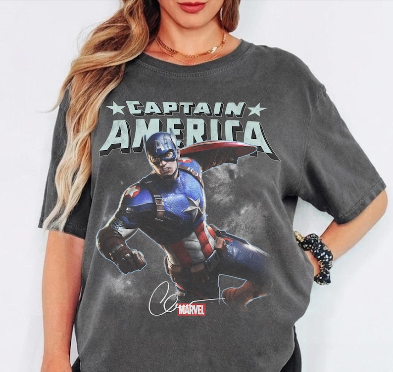 Puede incluir: Una camiseta de manga corta gris carb&oacute;n con un estampado gr&aacute;fico de Capit&aacute;n Am&eacute;rica en una pose din&aacute;mica. El texto CAPTAIN AMERICA est&aacute; impreso en una fuente clara y llamativa sobre el personaje, con un peque&ntilde;o logotipo rojo de Marvel abajo.