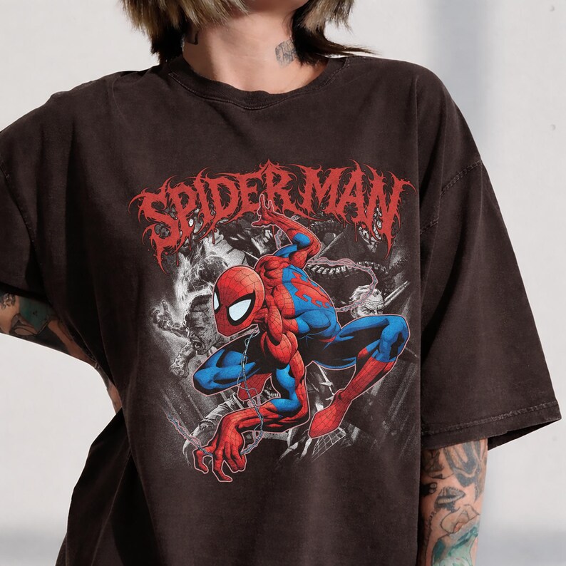 May include: A dark brown short-sleeve t-shirt featuring a graphic print of the comic book character Spider-Man in a crouching pose. The text SPIDER-MAN is printed in a stylized red font above the character, with black and white background imagery.