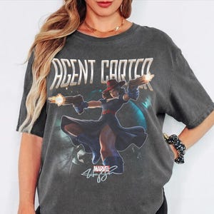 Puede incluir: Una camiseta gris carb&oacute;n de manga corta con un gr&aacute;fico de Agent Carter con vestido azul y sombrero rojo, sosteniendo dos pistolas disparando. El texto AGENT CARTER aparece en letras blancas grandes arriba, con un logo de Marvel y una firma.