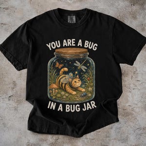 Op de afbeelding: Een zwart t-shirt met korte mouwen en een afbeelding van een kat met bijenvleugels in een glazen pot. De tekst luidt YOU ARE A BUG IN A BUG JAR. De illustratie bevat een vlinder, een libel, paddenstoelen en kleine bloemen in de pot.