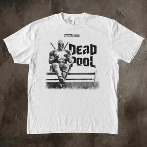 May include: A white short-sleeve t-shirt featuring a black and white graphic of the character Deadpool sitting and reading a paper. The text MARVEL STUDIOS is printed above the character, and the word DEADPOOL is printed in large letters to the right.