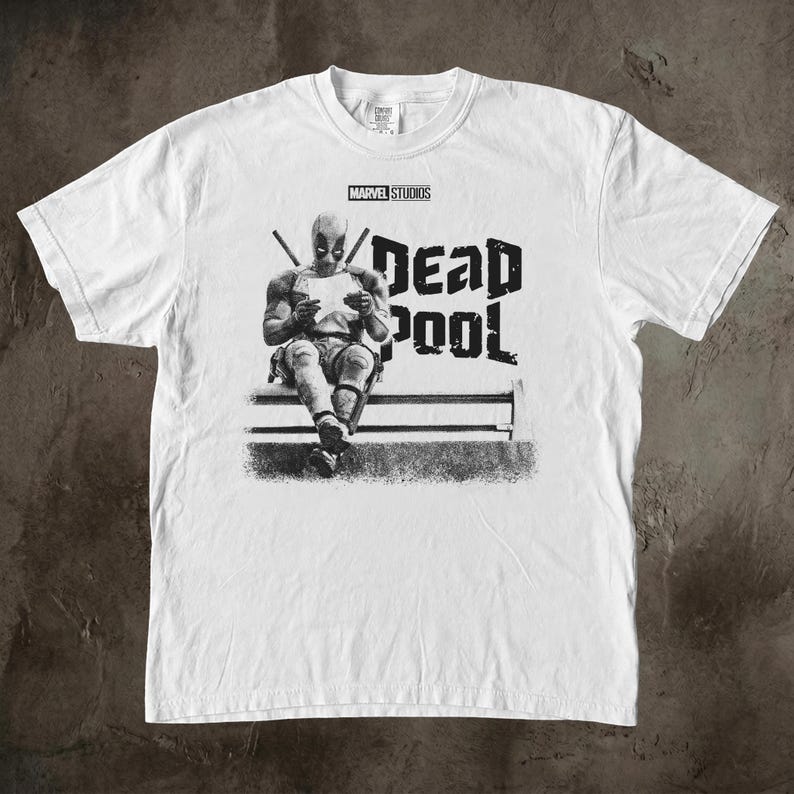 May include: A white short-sleeve t-shirt featuring a black and white graphic of the character Deadpool sitting and reading a paper. The text MARVEL STUDIOS is printed above the character, and the word DEADPOOL is printed in large letters to the right.