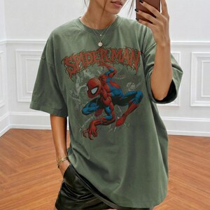 May include: An olive green short-sleeve t-shirt featuring a graphic print of the comic book character Spider-Man in a crouching pose. The text SPIDER-MAN is printed in a distressed red font above the character. The shirt has a crew neckline.