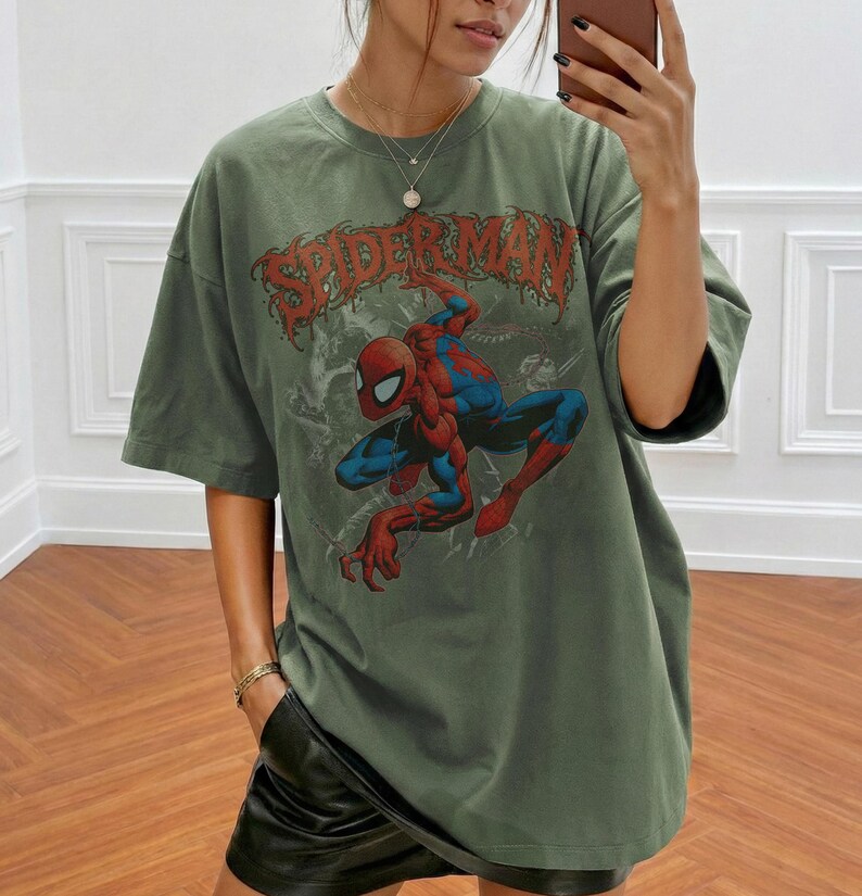 May include: An olive green short-sleeve t-shirt featuring a graphic print of the comic book character Spider-Man in a crouching pose. The text SPIDER-MAN is printed in a distressed red font above the character. The shirt has a crew neckline.