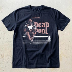 May include: A dark navy blue short-sleeve t-shirt featuring a graphic print of the character Deadpool sitting on a ledge while reading a letter. The text "MARVEL STUDIOS" appears above the character, and the word "DEADPOOL" is printed in large letters.