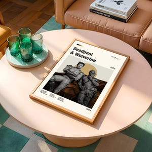 May include: A framed poster featuring Deadpool and Wolverine. The poster has a light beige background with the title "Deadpool & Wolverine" in bold black text. The image shows the two characters in a sepia tone. The frame is made of light wood.