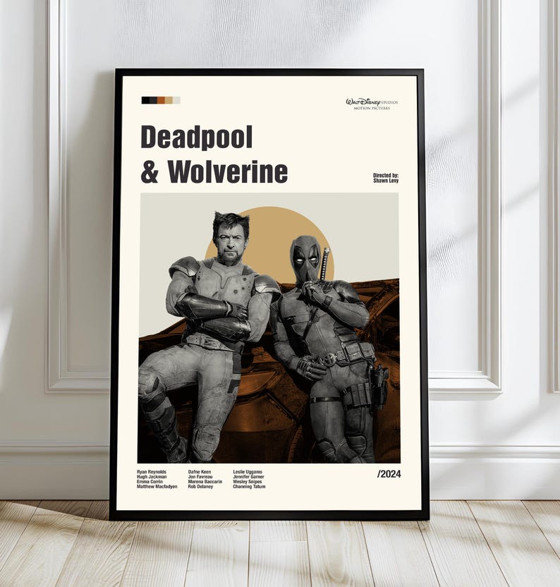 May include: A framed poster featuring Deadpool and Wolverine. The poster has a beige background with the title "Deadpool & Wolverine" in bold black letters. The image shows the two characters in costume, with a sun-like shape behind them. The poster is framed in black.