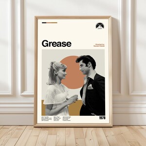 Può includere: Un poster incorniciato del film "Grease" con uno sfondo beige e il titolo in nero. Il poster mostra i due personaggi principali, una donna con un vestito bianco e un uomo con un abito nero. L'anno 1978 è visualizzato. Il poster è in una cornice di legno.