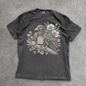 May include: A charcoal grey short-sleeve t-shirt featuring a detailed cream-coloured graphic print of a crow perched on a branch. The crow is depicted knitting with two needles and a ball of yarn, surrounded by floral elements and leaves.