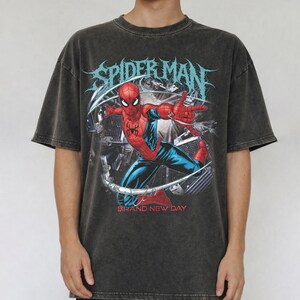 May include: A charcoal gray, acid-washed short-sleeve t-shirt featuring a graphic print of Spider-Man in a dynamic pose. The text "SPIDER-MAN" appears in a stylized font above the character, with the words "BRAND NEW DAY" printed in red below.