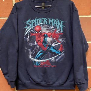 May include: A navy blue crewneck sweatshirt featuring a graphic print of Spider-Man in a dynamic pose. The design includes the text "SPIDER-MAN" in a stylized font at the top and "BRAND NEW DAY" in red lettering at the bottom of the illustration.