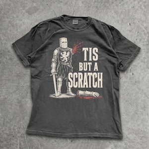 May include: A charcoal grey short-sleeve t-shirt featuring a graphic of a medieval knight with a severed arm. The knight holds a sword and stands near the detached arm. The text "TIS BUT A SCRATCH" is printed in large white capital letters.