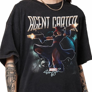 Puede incluir: Una camiseta negra de manga corta con un estampado gr&aacute;fico de Agent Carter sosteniendo dos pistolas con destellos de disparo. El texto AGENT CARTER aparece en letras blancas grandes en la parte superior, con un logo de Marvel y una firma abajo.