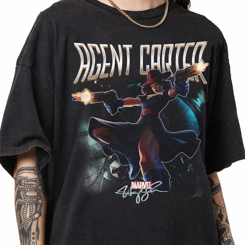Puede incluir: Una camiseta negra de manga corta con un estampado gr&aacute;fico de Agent Carter sosteniendo dos pistolas con destellos de disparo. El texto AGENT CARTER aparece en letras blancas grandes en la parte superior, con un logo de Marvel y una firma abajo.