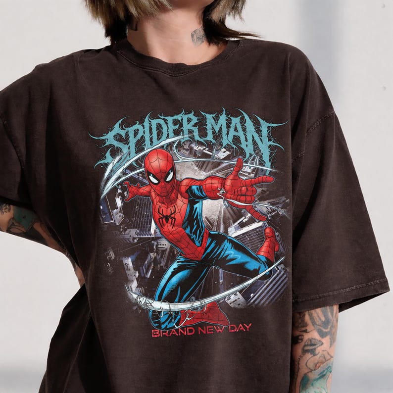 May include: A dark brown short-sleeve t-shirt featuring a graphic print of Spider-Man in a dynamic pose. The design includes the text "SPIDER-MAN" in a stylized font at the top and "BRAND NEW DAY" in red lettering at the bottom, set against a city background.