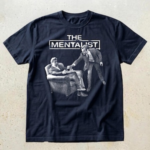 May include: A dark navy blue short-sleeve t-shirt featuring a white graphic print of two figures in suits. One figure sits in an armchair while the other stands, with the text THE MENTALIST printed in a white rectangular frame above the figures.