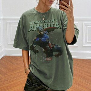 Puede incluir: Una camiseta de corte oversize en color verde oliva con un estampado gr&aacute;fico de Capit&aacute;n Am&eacute;rica en una pose din&aacute;mica. El texto CAPTAIN AMERICA aparece en letras grandes de color verde claro sobre el personaje, con un logo rojo de Marvel abajo.