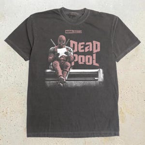 May include: A charcoal gray short-sleeve t-shirt featuring a graphic of the character Deadpool sitting and reading a paper. The text MARVEL STUDIOS appears above the character, and the word DEADPOOL is printed in large, distressed pink letters.