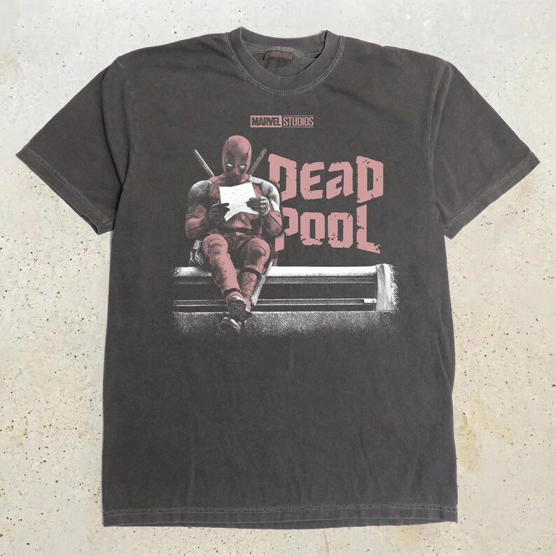 May include: A charcoal gray short-sleeve t-shirt featuring a graphic of the character Deadpool sitting and reading a paper. The text MARVEL STUDIOS appears above the character, and the word DEADPOOL is printed in large, distressed pink letters.